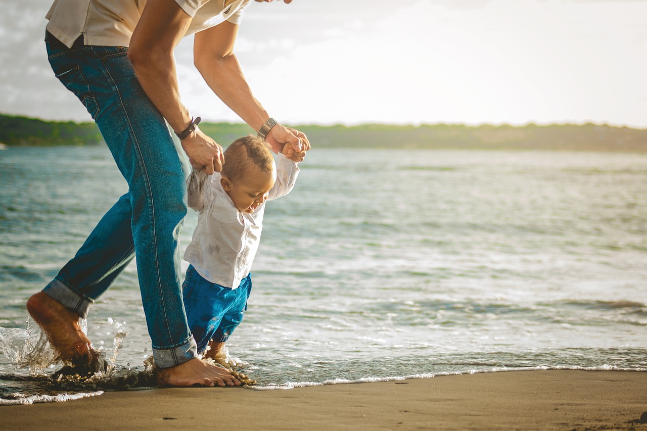 Szene am Meer, Strand: Vater hält Hände seines Babys, das versucht zu gehen.